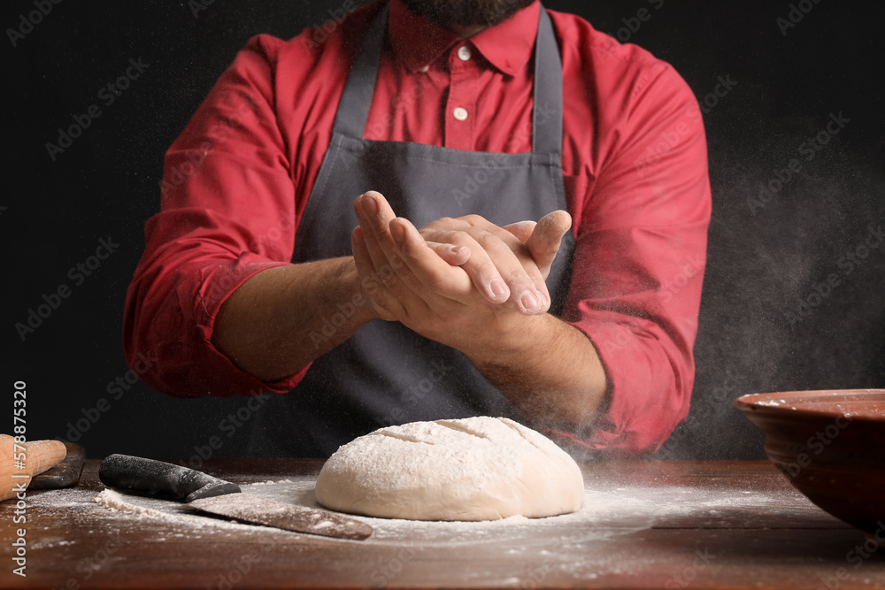 Male baker sprinkling dough with flour at table on dark background, closeup