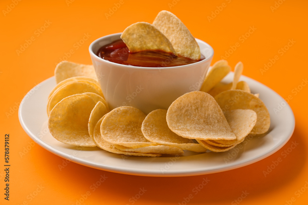 Plate with delicious potato chips and bowl of ketchup on orange background