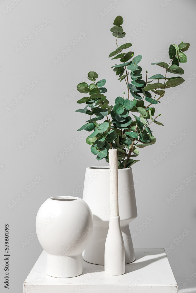 Vases with eucalyptus branches and candle on table near grey wall