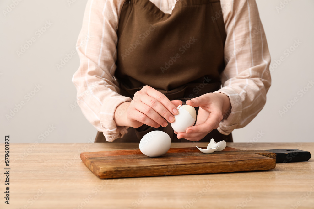 Woman peeling boiled egg at table near grey wall