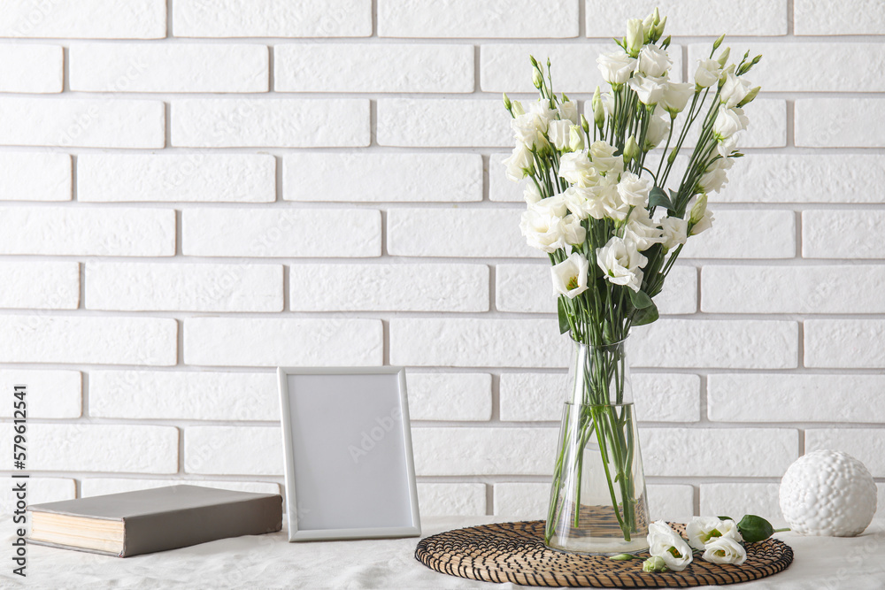 Vase with beautiful eustoma flowers, book and blank picture frame on table near light brick wall