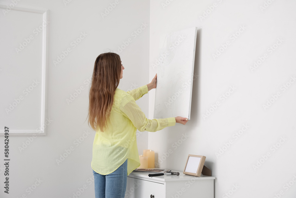 Young woman hanging painting on light wall at home