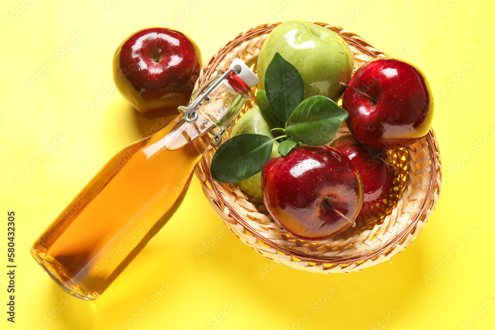 Glass bottle of fresh apple cider vinegar and basket with fruits on yellow background