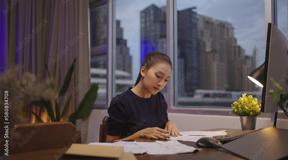 Asian woman sits at her desk with paperwork and computer at home working late at night.