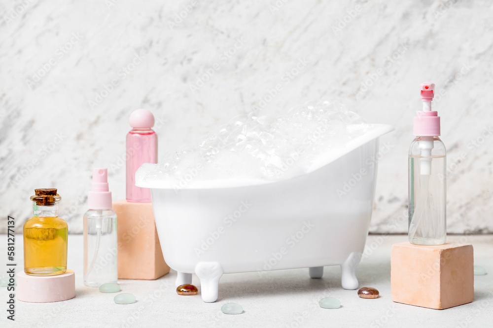 Small bathtub with foam and bottles of cosmetic products on light background