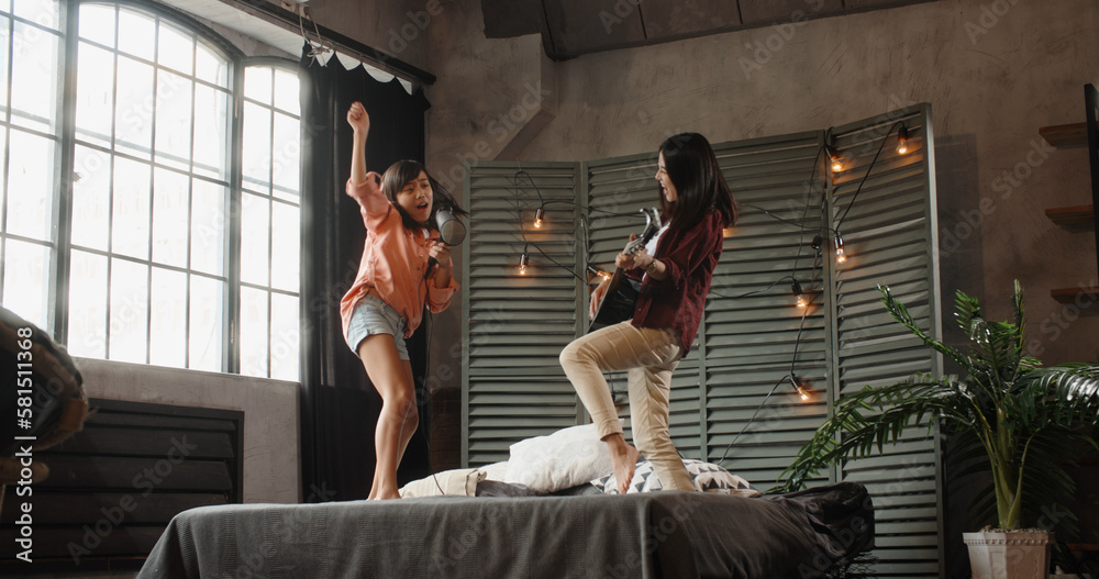 Asian sisters are having fun at home, one is playing guitar, other sings into hairdryer while jumpin