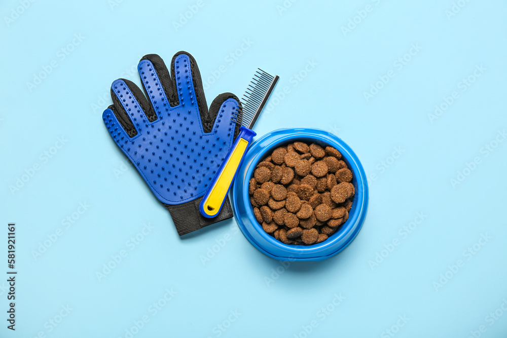 Bowl of dry pet food, grooming glove and comb on color background