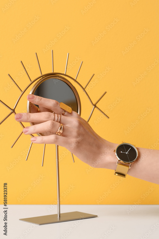 Woman with golden wristwatch and mirror on table near yellow wall