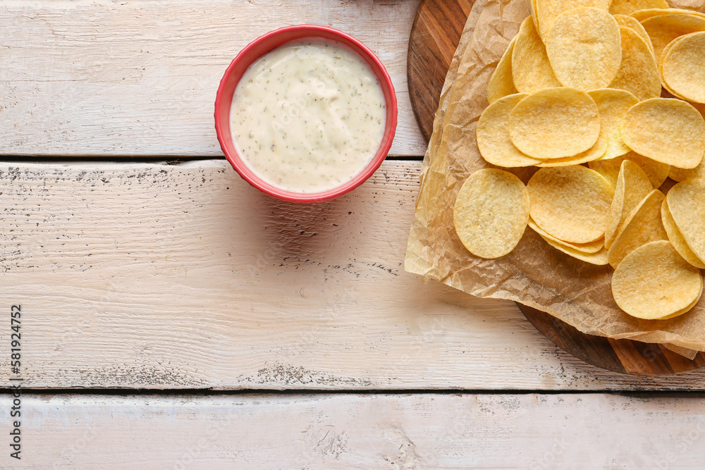 Board with delicious potato chips and bowl of sauce on white wooden background