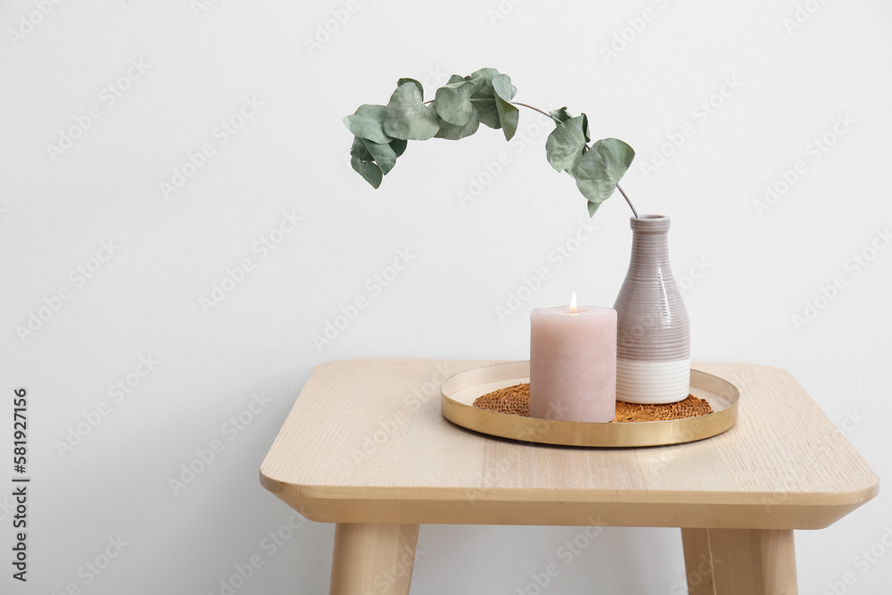 Burning candle and vase with eucalyptus branch on table near light wall