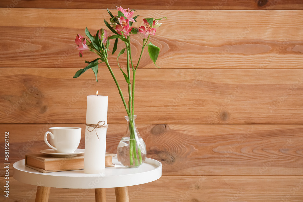 Burning candle, cup of coffee, book and vase with flowers on table near wooden wall