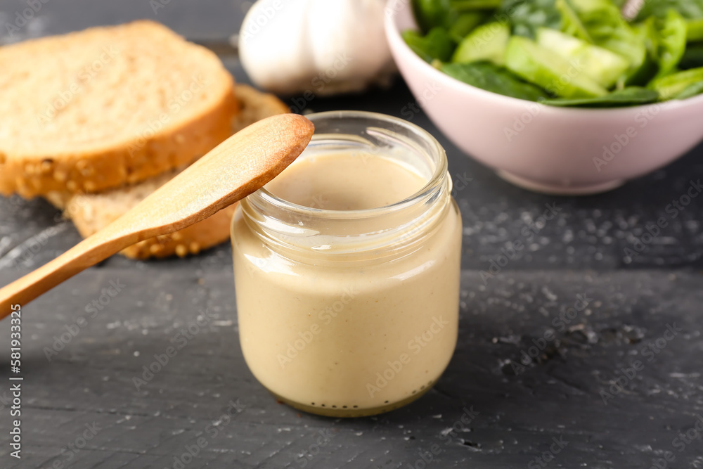 Glass jar of tasty tahini, spoon, bread and salad on dark wooden table