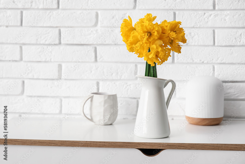 Vase with narcissus flowers and cup of tea on table near white brick wall