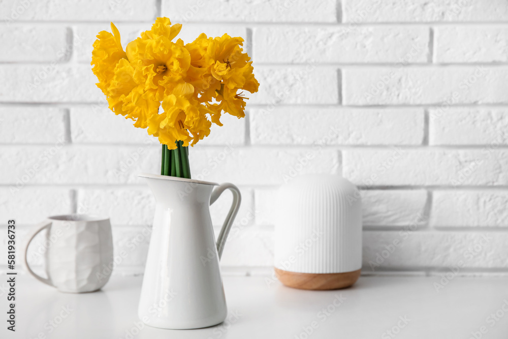 Vase with narcissus flowers and cup of tea on table near white brick wall