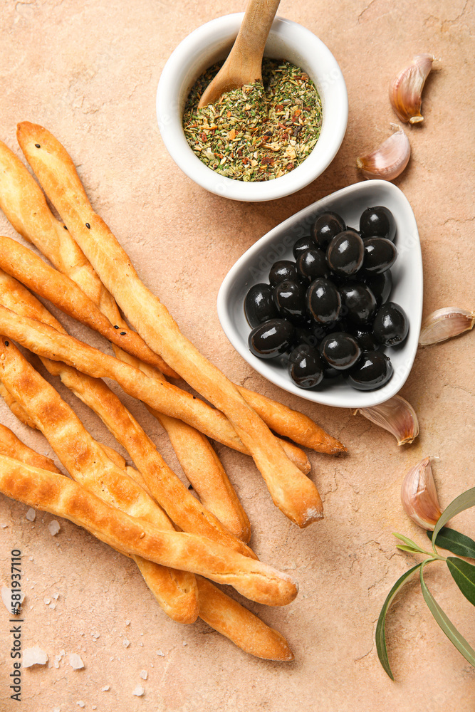 Tasty Italian Grissini with bowls of herbs and black olives on beige background