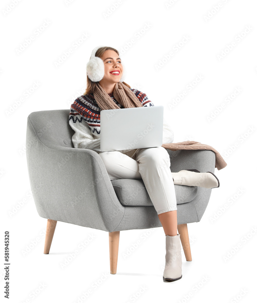 Young woman with earmuffs using laptop in grey armchair on white background