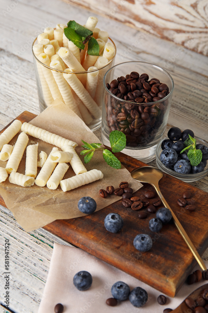 Board with delicious wafer rolls, blueberries and coffee beans on white wooden background