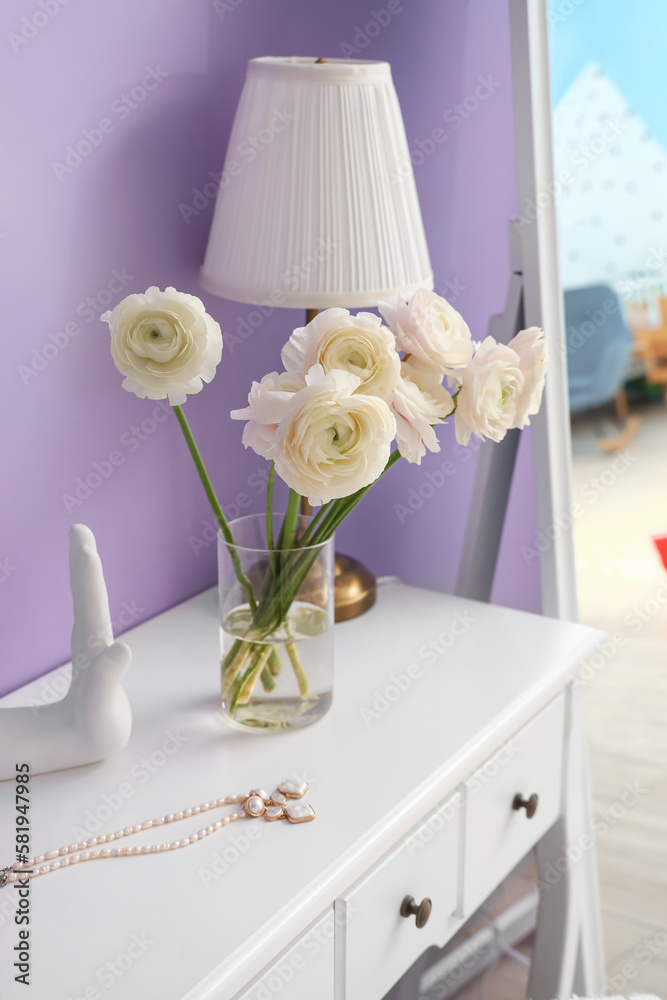 Vase with ranunculus flowers, jewelry and lamp on dressing table in room