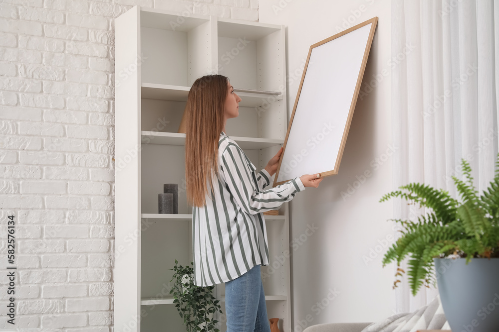 Young woman hanging blank frame on white brick wall in bathroom