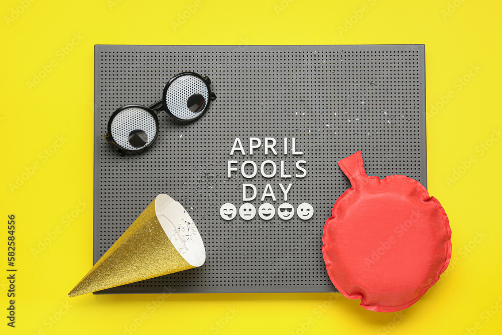 Board with text APRIL FOOLS DAY, funny decor and party hat on yellow background