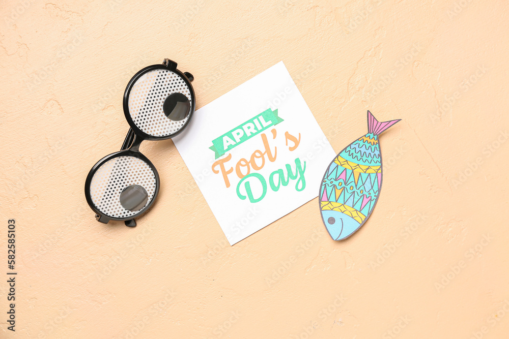 Paper with text APRIL FOOLS DAY, fish and funny eyeglasses on beige background