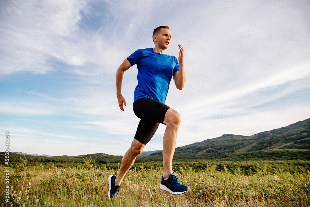 sporty male runner running on mountain trail in summer, front view, blue shirt and black half tights