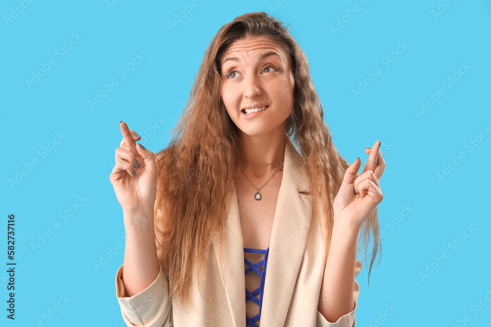 Beautiful young woman in jacket crossing fingers on blue background