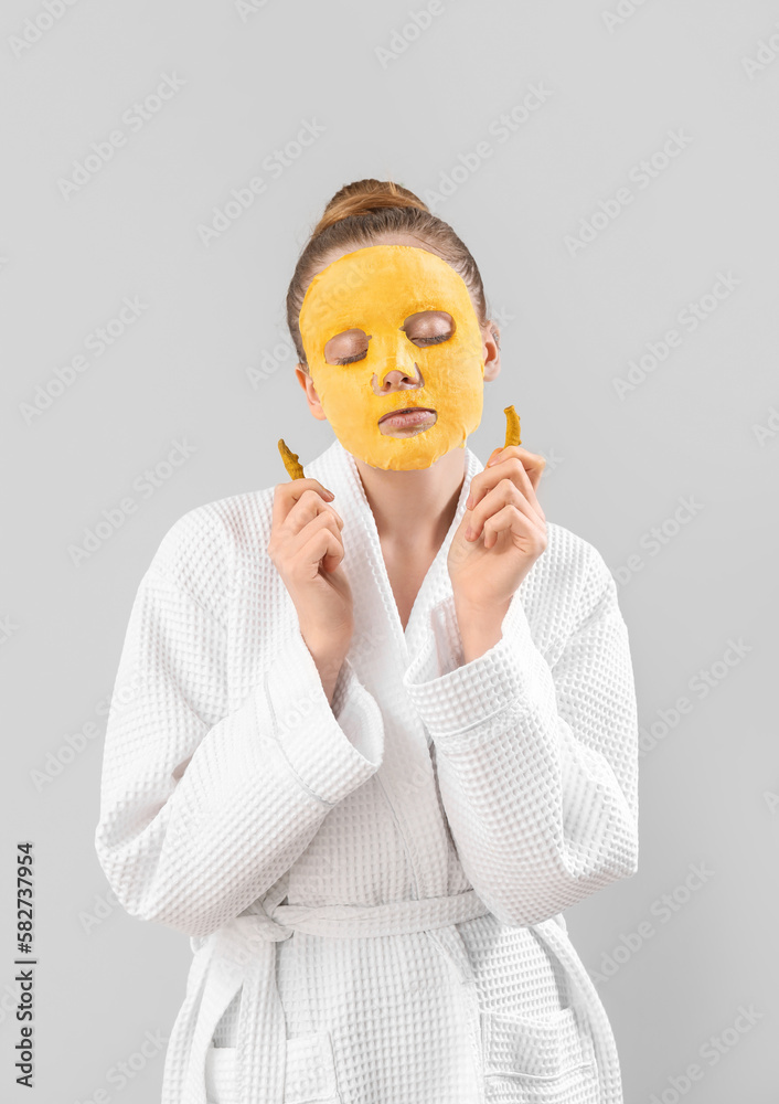 Young woman in bathrobe with turmeric sheet mask and roots on grey background