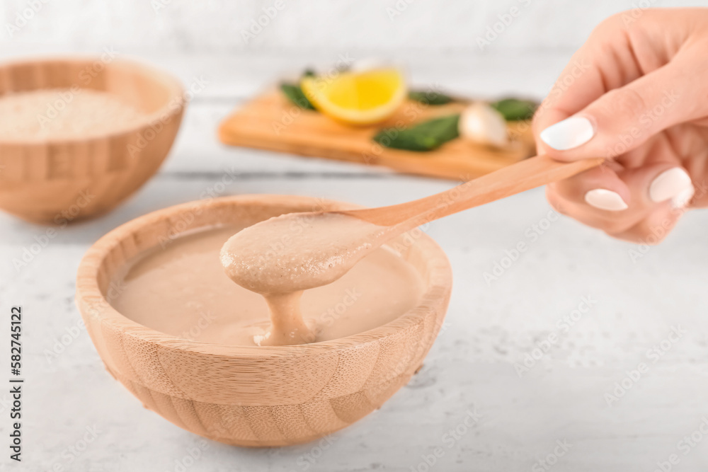 Female hand with spoon and bowl of tasty tahini on light wooden table, closeup