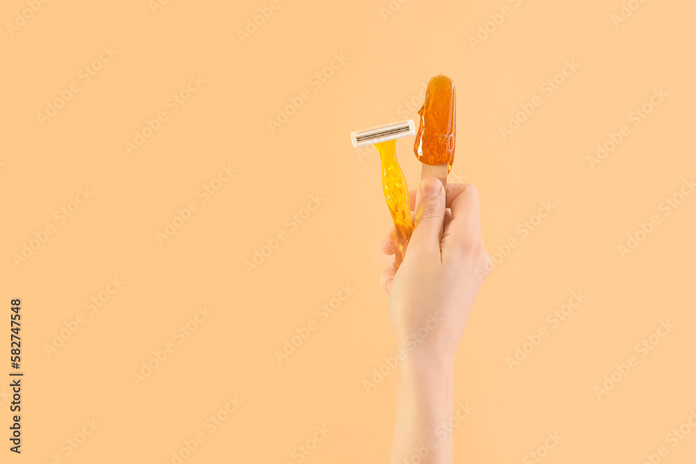 Female hand holding spatula with sugaring paste and razor on beige background