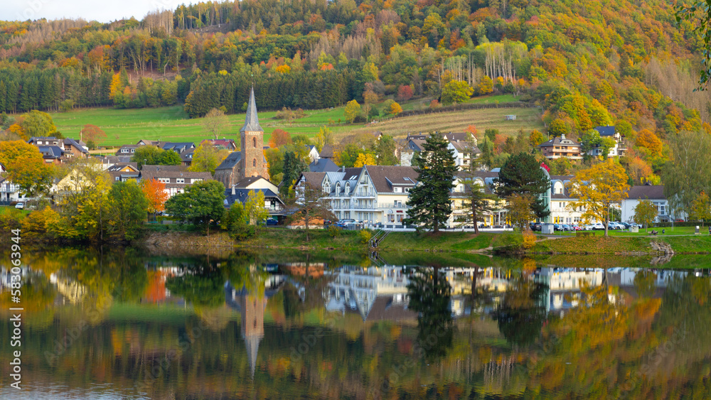 Beautiful german village with colorful foliage forest in the background and reflection in the Rursee