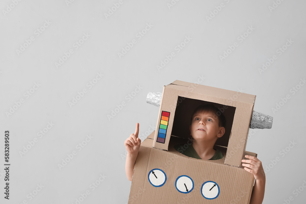 Little boy in cardboard robot costume pointing at something near light wall