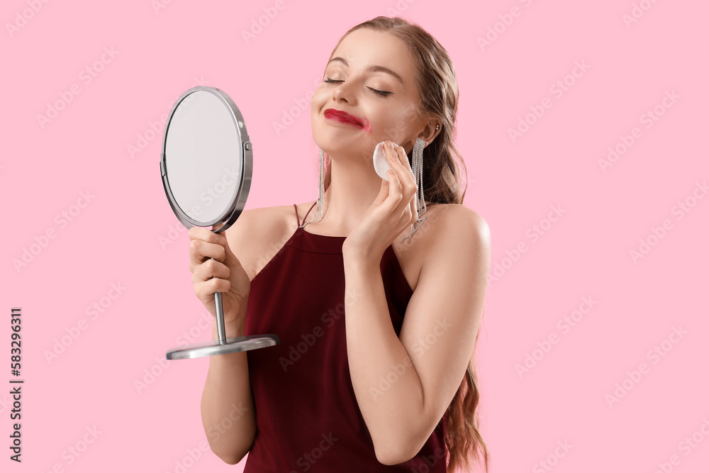 Young woman with mirror removing makeup on pink background