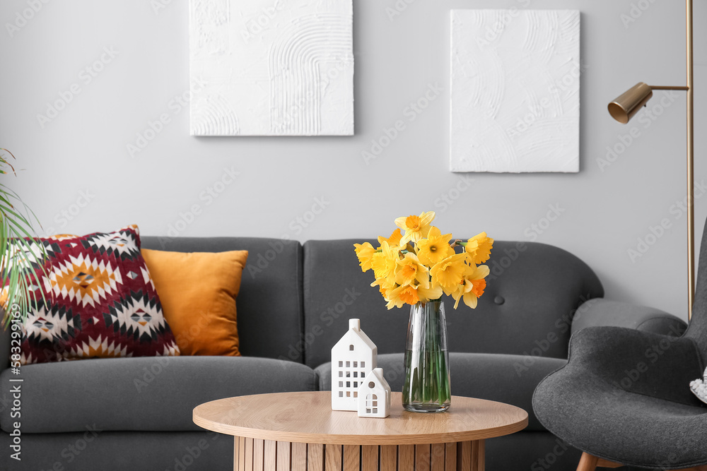 Porcelain houses and vase with narcissus flowers on coffee table near sofa in living room