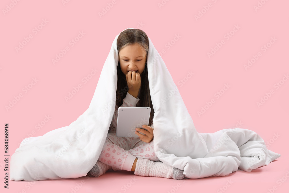 Little girl with blanket and tablet computer biting nails on pink background