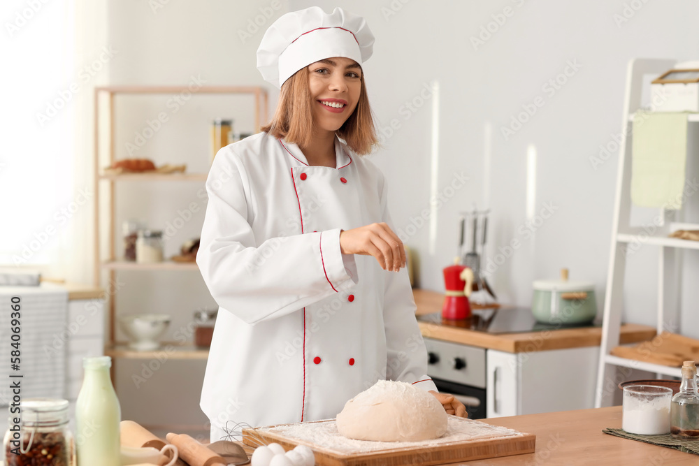 Female baker sprinkling dough with flour at table in kitchen