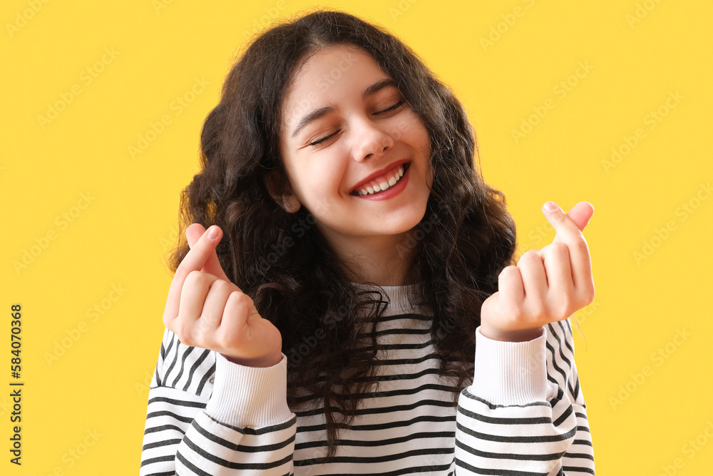 Teenage girl making heart shape with her fingers on yellow background, closeup