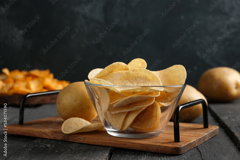 Board with bowl of delicious potato chips on wooden table against black background