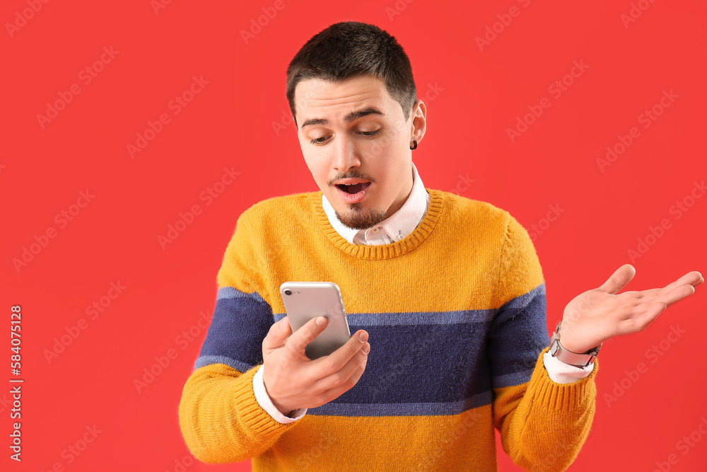 Confused young man with smartphone on red background