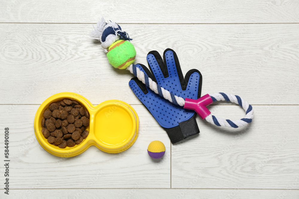 Bowl of dry pet food, toys and grooming glove on light wooden background