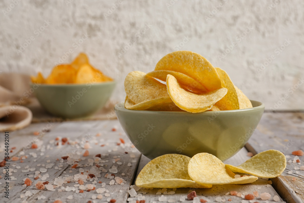 Bowl with delicious potato chips and sea salt on wooden table against grey background, closeup