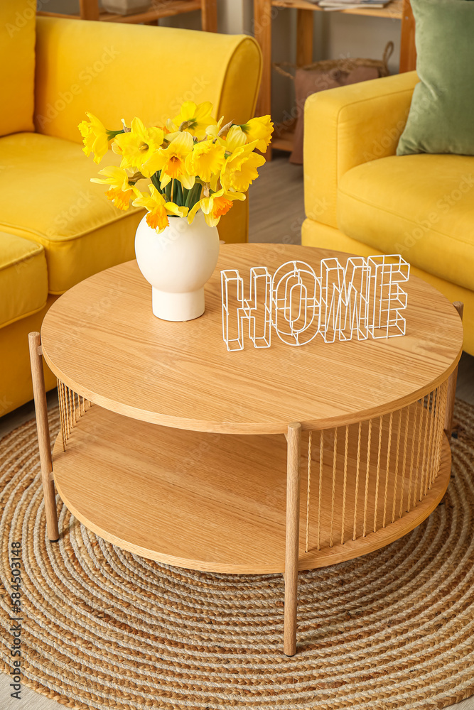 Vase with narcissus flowers and word HOME on coffee table in living room