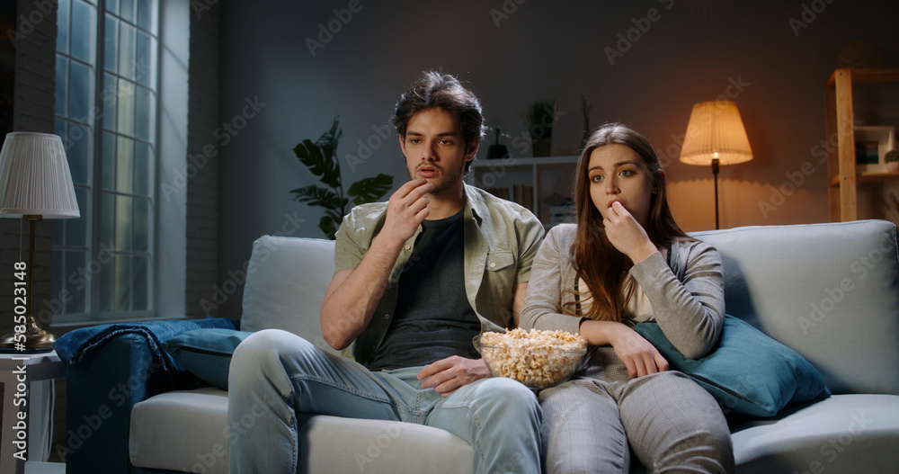 Young student caucasian concentrated couple sitting on couch in the evening watching tv show and eat