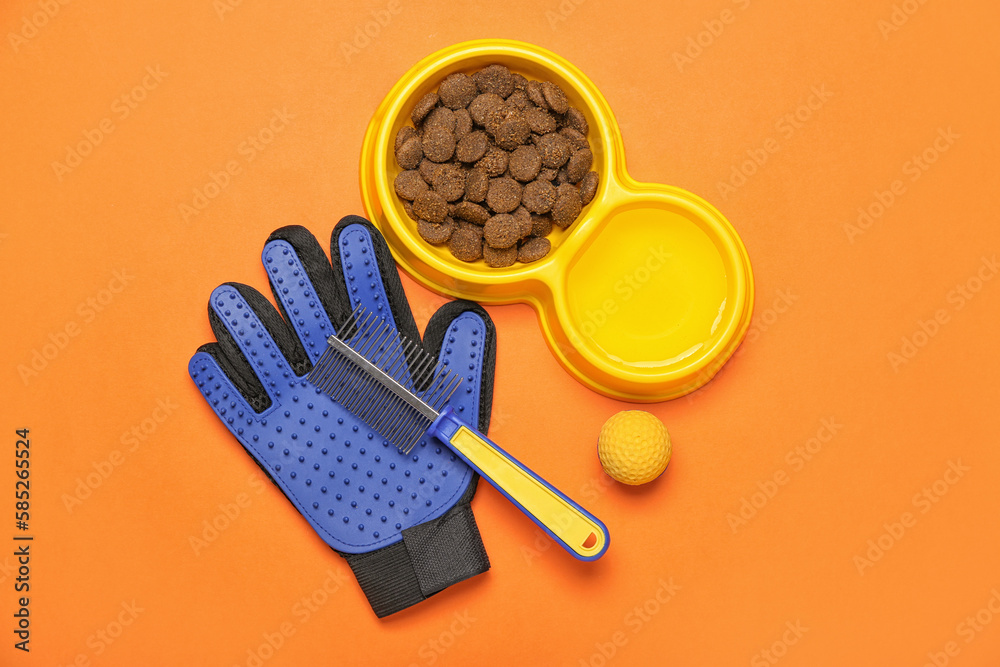 Bowl of dry pet food, rubber ball, grooming glove and comb on color background