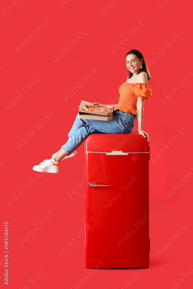 Beautiful woman with tasty pizza sitting on fridge against red background