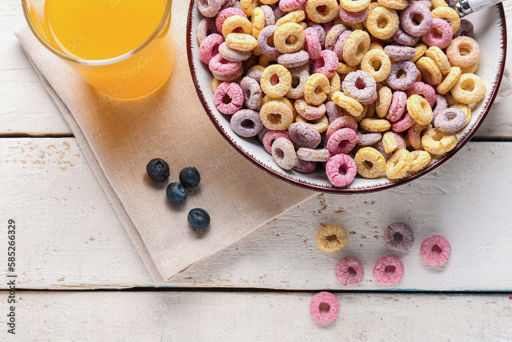 Bowl with colorful cereal rings, glass of juice and blueberries on white wooden background