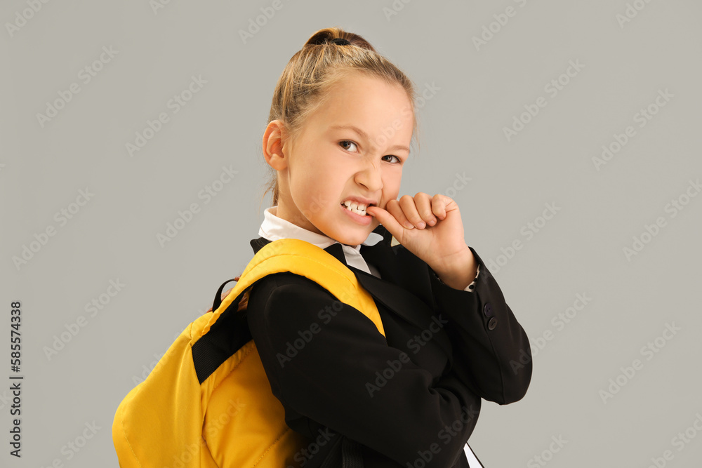 Little schoolgirl with backpack biting nails on grey background