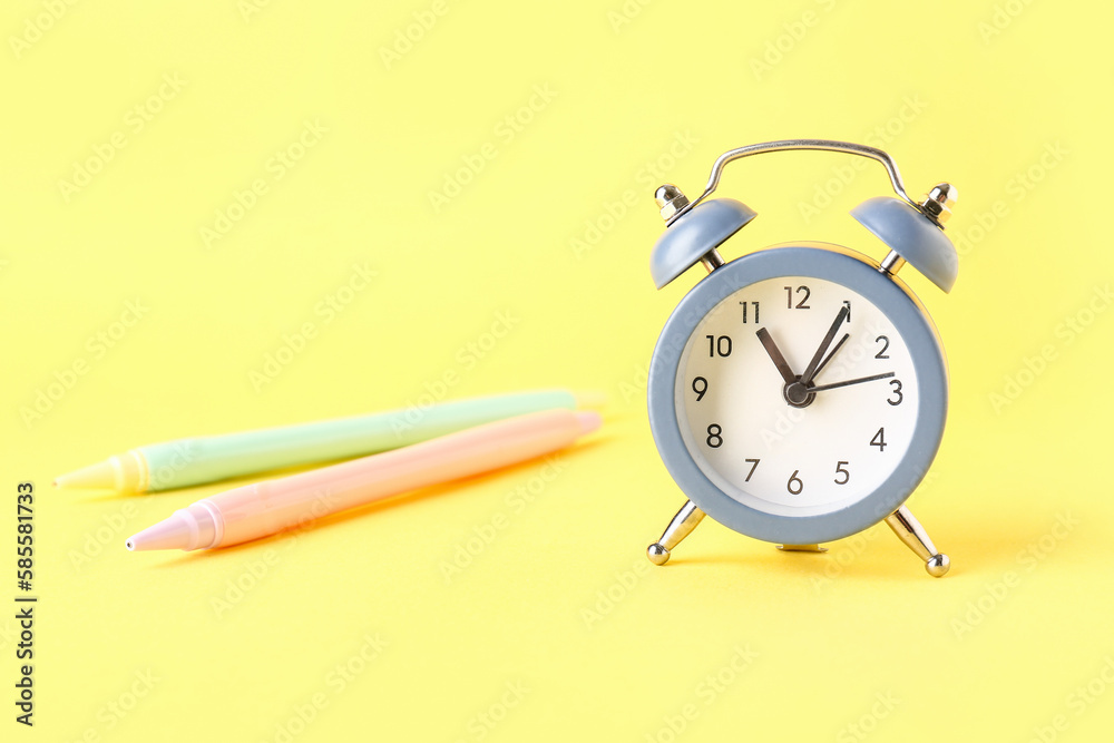 Alarm clock and pens on yellow background