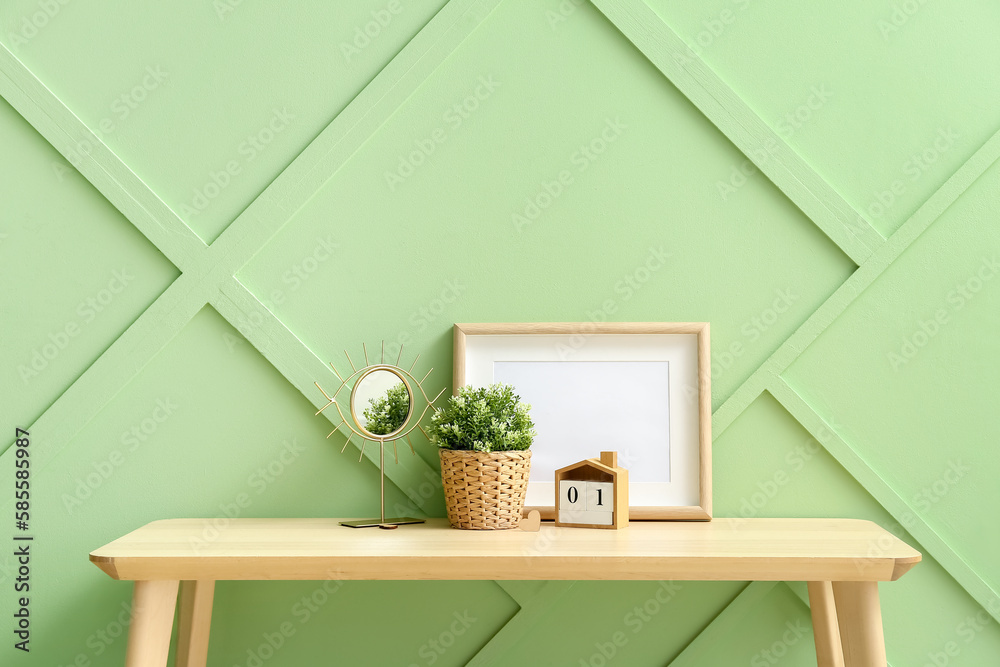 Artificial houseplant with mirror, calendar and blank frame on table near green wall