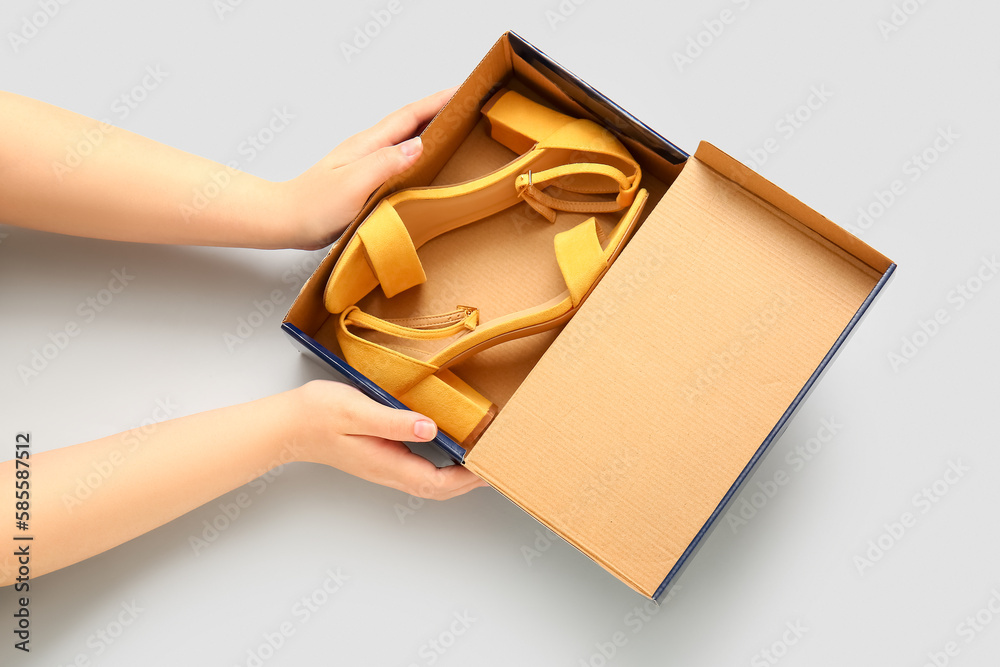 Female hands holding cardboard box with stylish shoes on light background